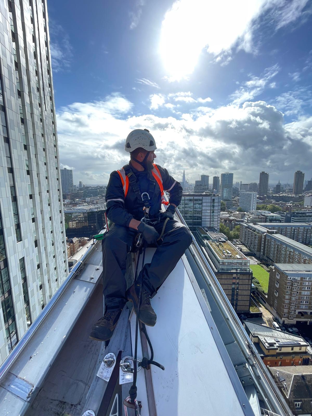 Técnico com EPI completo sentado no bordo da cobertura de um edifício alto, com as cordas montadas